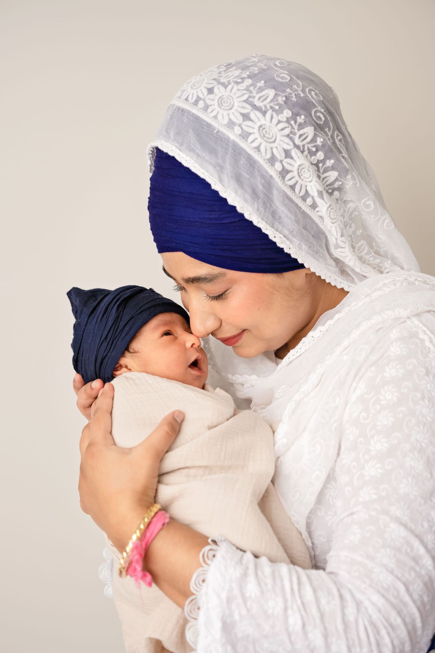 Sikh mother in white embroidered dupatta and blue turban tenderly holds her newborn baby wrapped in matching blue headwrap.