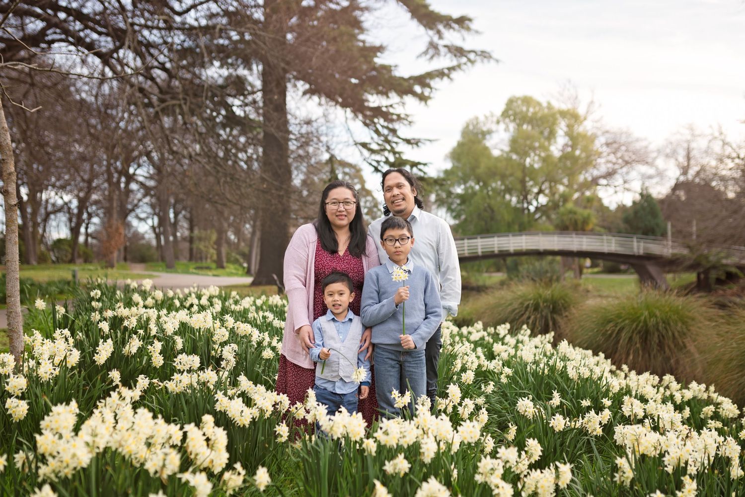 Family of four posing among white daffodils in a lush park with trees and a bridge in the background.