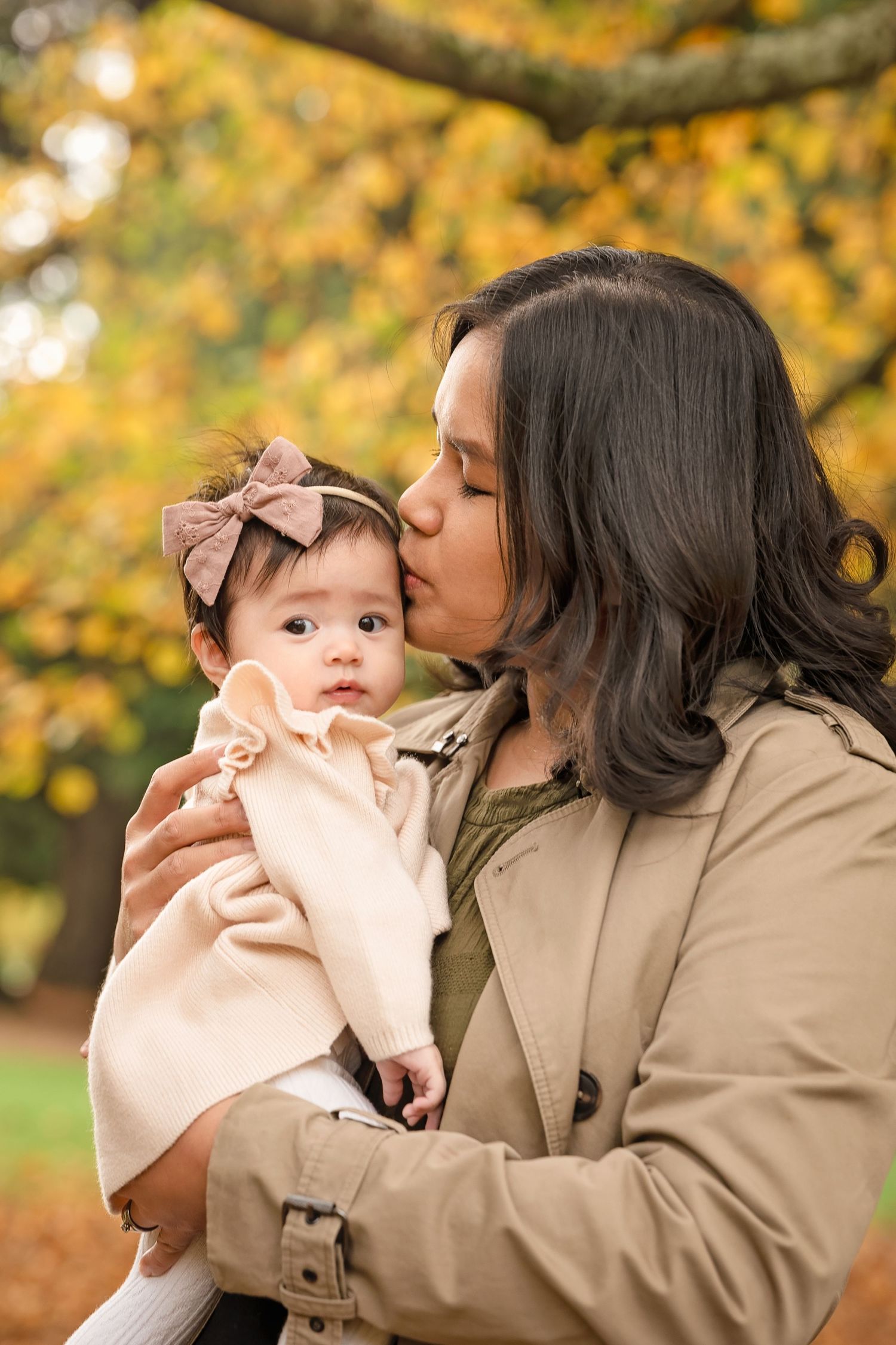 Mother kissing baby girl with bow headband outdoors in autumn park with golden yellow foliage background.