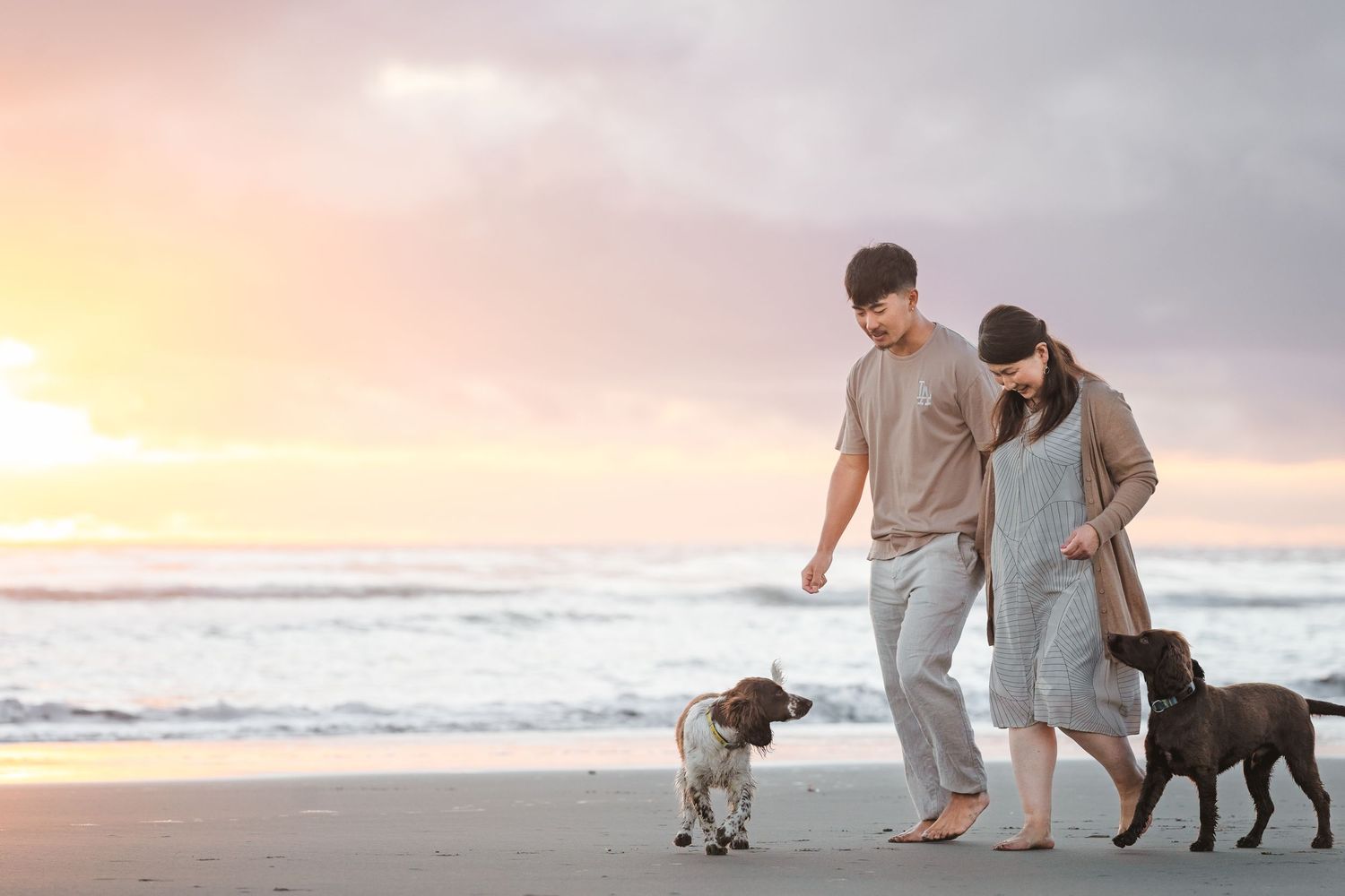 Couple walking their two dogs on a beach at sunset with gentle waves rolling in.