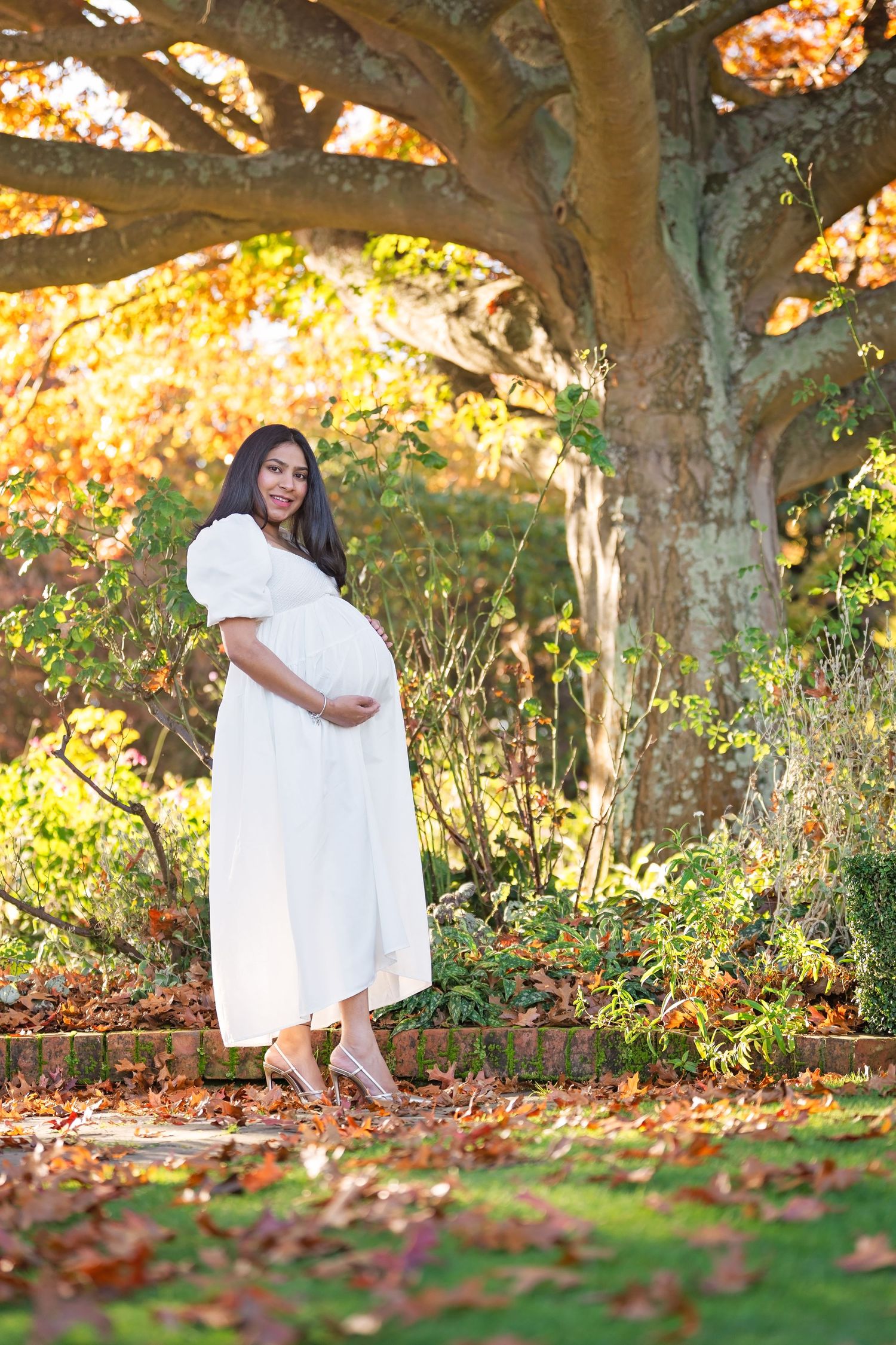 A pregnant figure in a flowing white dress poses under a large autumn tree surrounded by golden falling leaves in a garden setting.