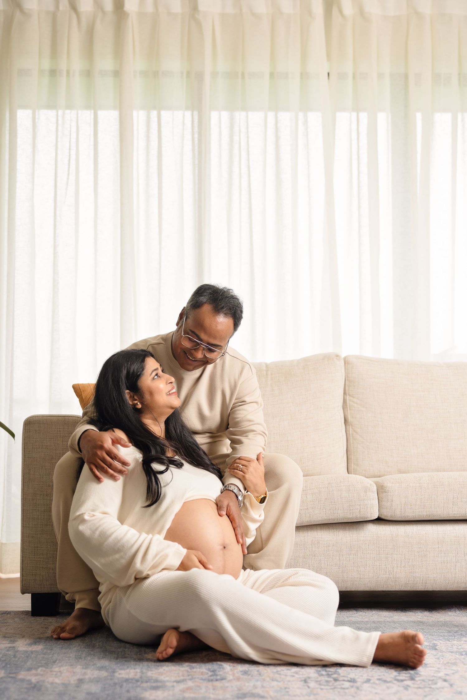 Two people wearing white loungewear relax intimately together on a beige couch in a bright sunlit room.