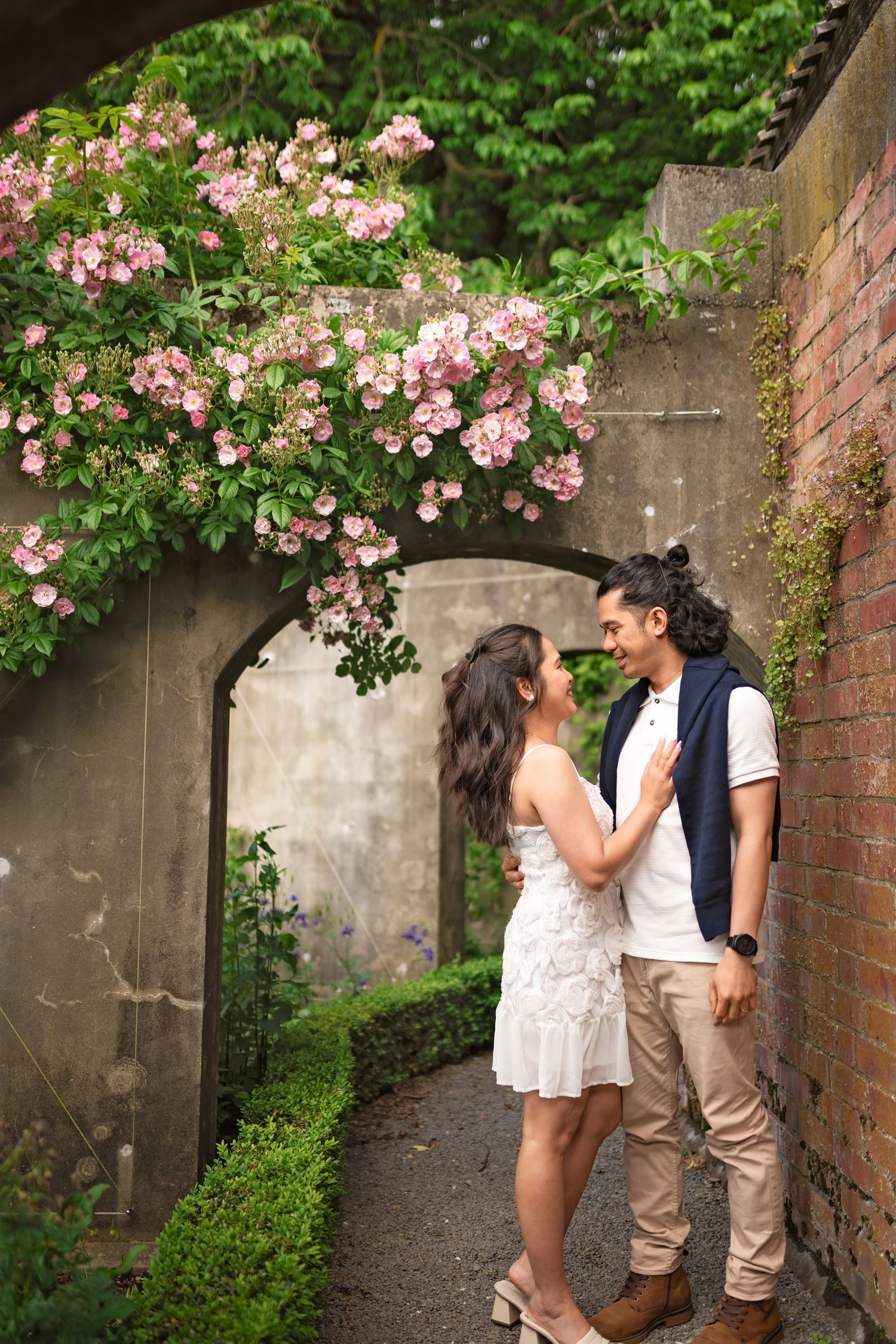 Romantic couple embracing under pink climbing roses in a charming garden archway with brick walls.