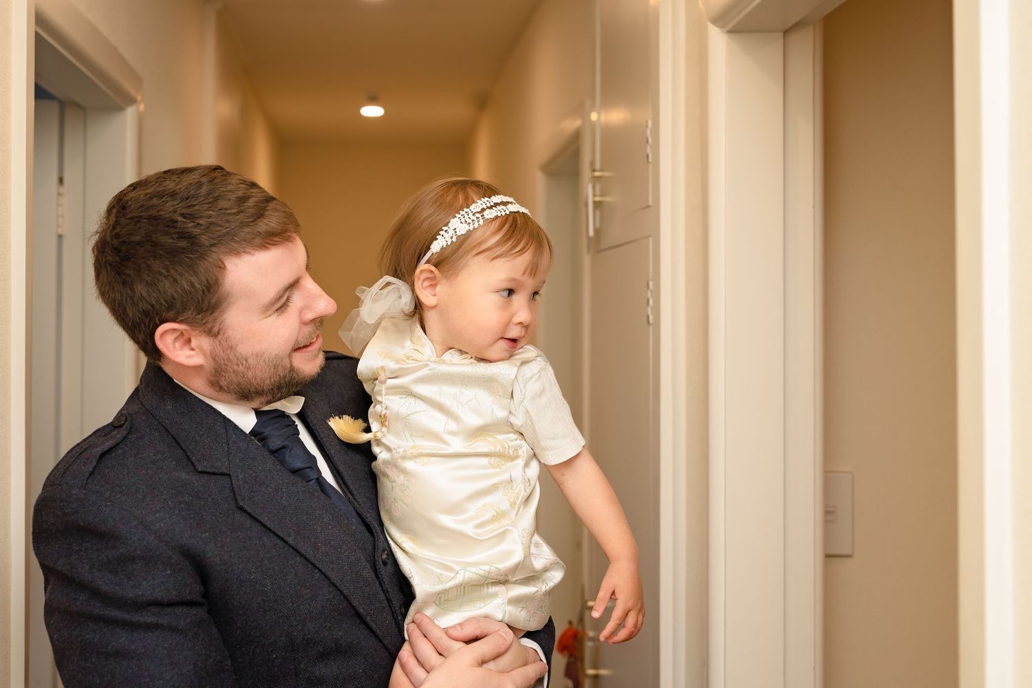 Smartly dressed groom in suit holds a young toddler in white outfit in a hotel corridor.