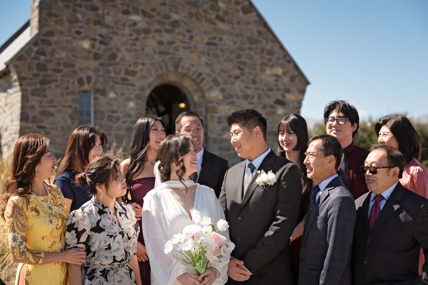 Wedding couple and guests gather outside a historic stone chapel under a bright blue sky for a group photo.