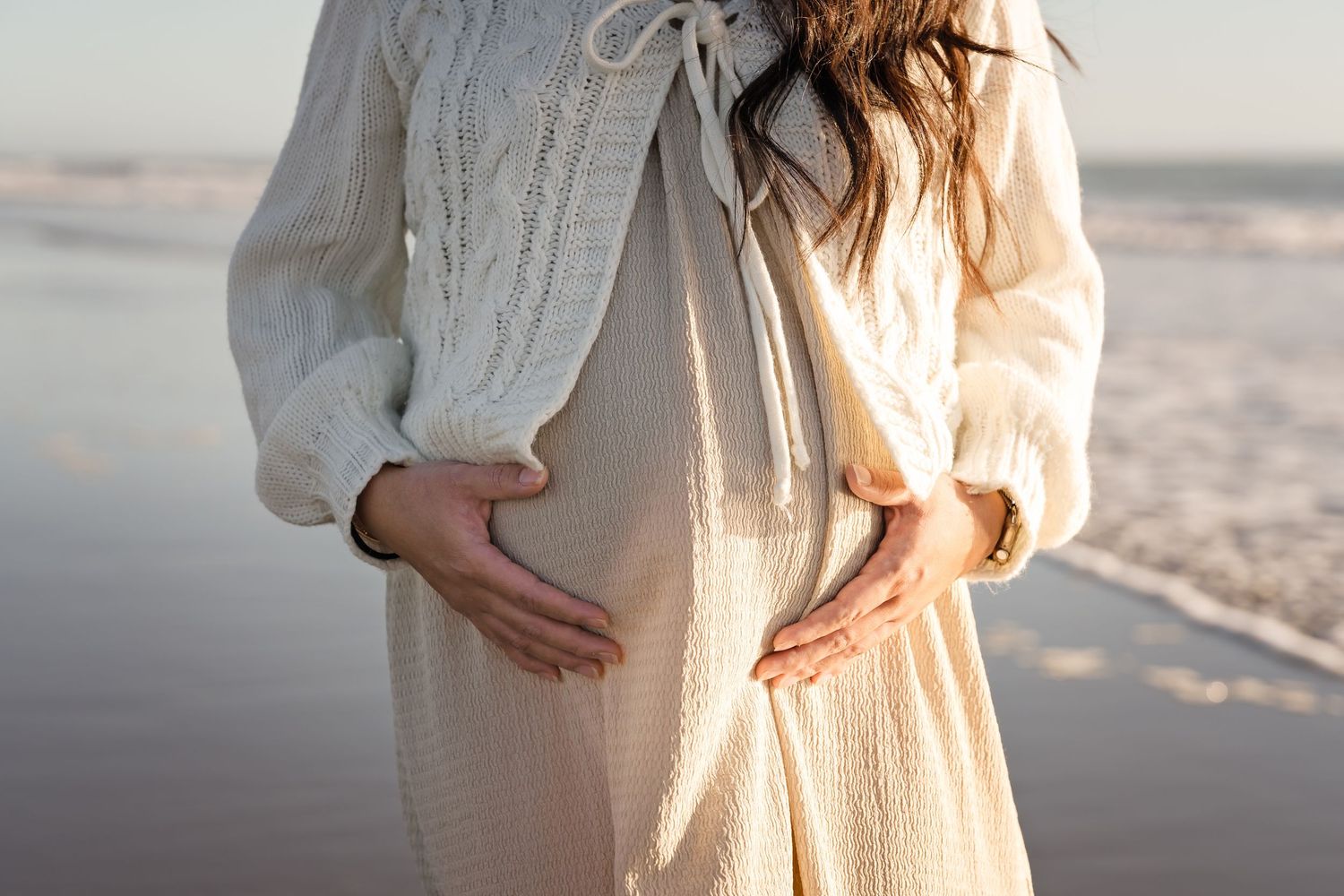 Pregnant woman in cream knit cardigan cradling baby bump on a serene beach, ocean waves in background.
