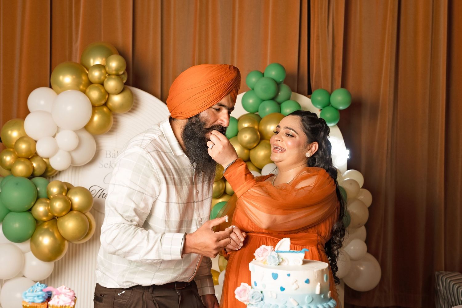 Sikh man feeds cake to smiling woman in orange dress at baby shower celebration with balloon decorations.