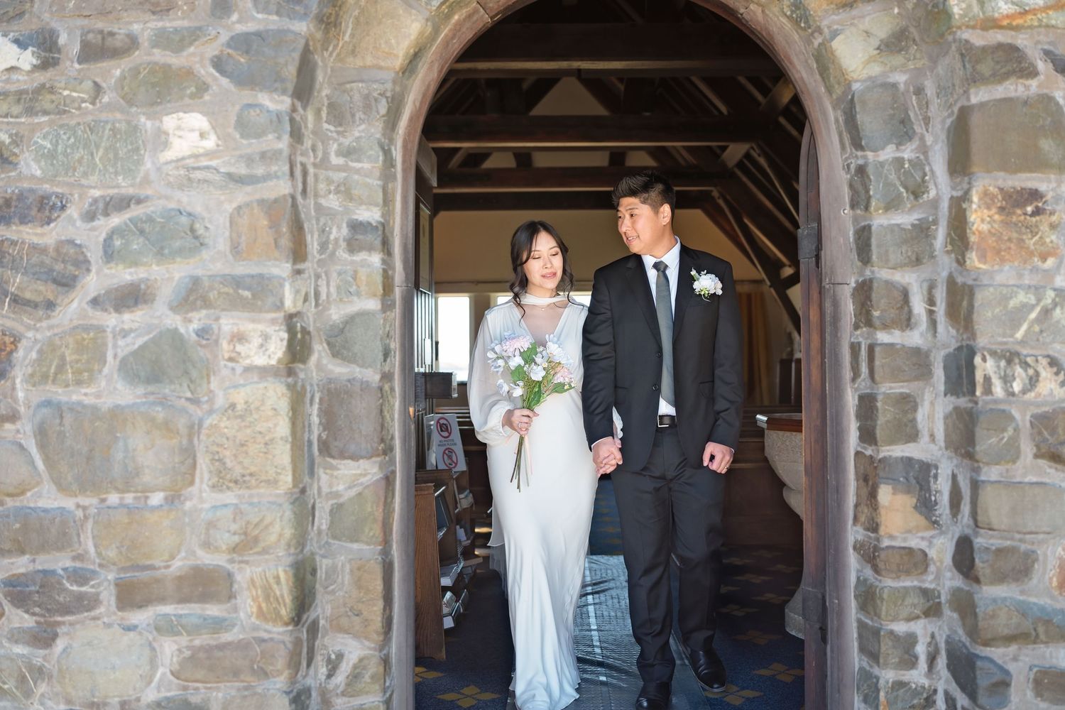 Bride and groom walk hand in hand through a stone archway after their wedding ceremony.