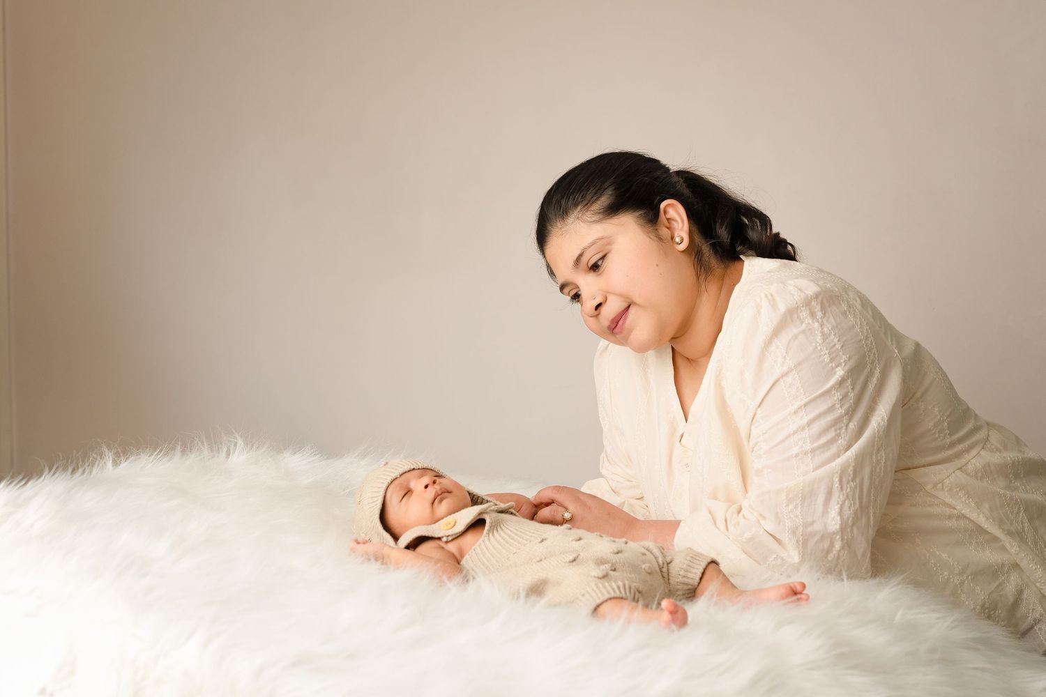 A mother tenderly gazes at her newborn baby lying on a white fluffy blanket during a professional newborn photo session.