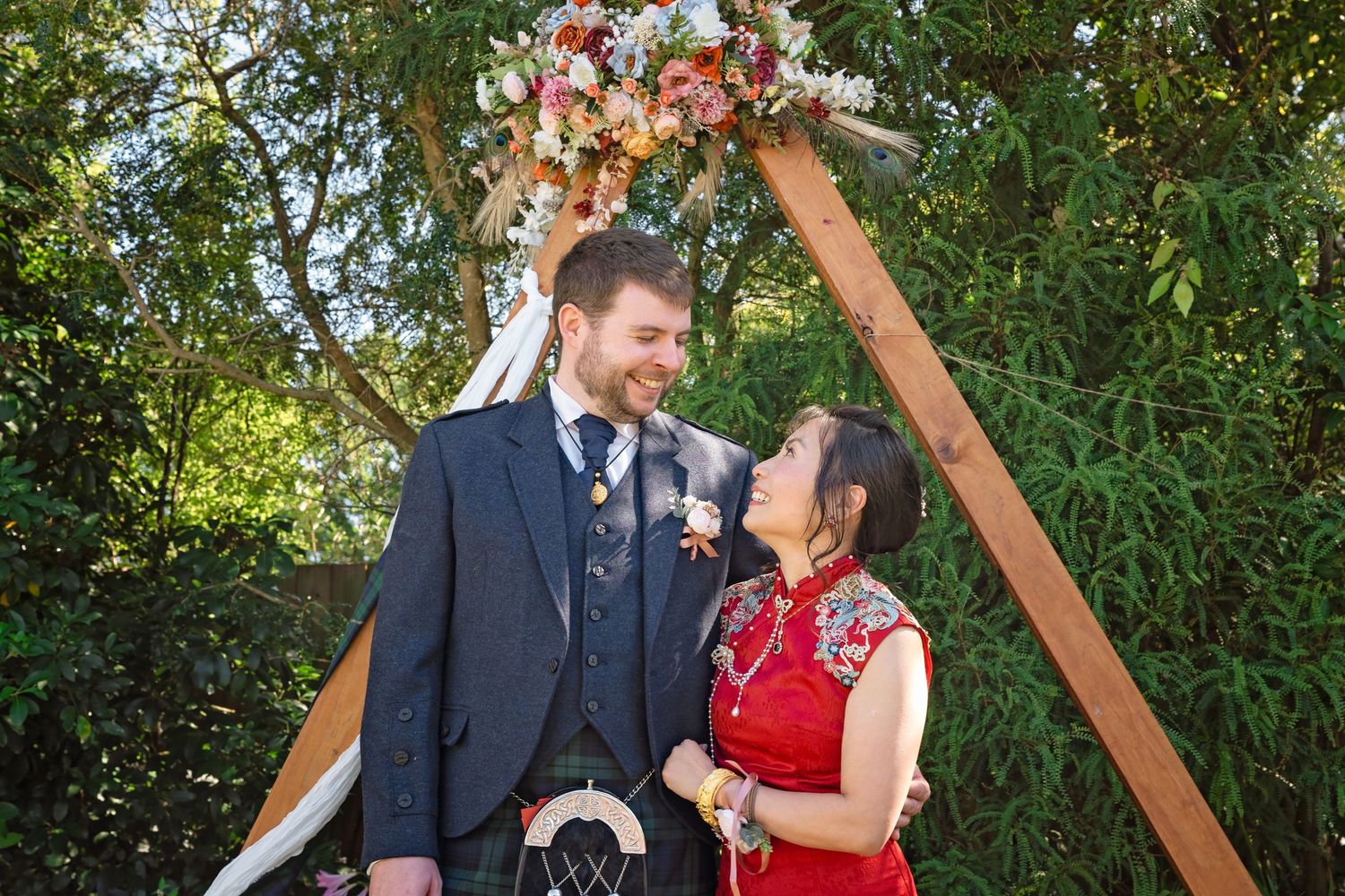 Bride in red qipao and groom in Scottish kilt share a romantic moment under floral wedding arch.