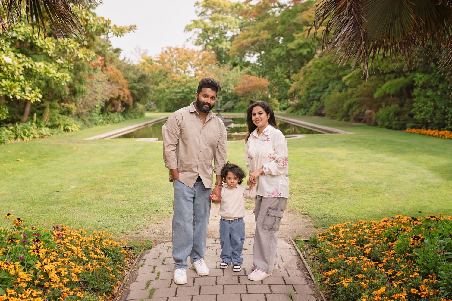 Family of three posing on a brick path surrounded by orange flowers and lush greenery in a park.