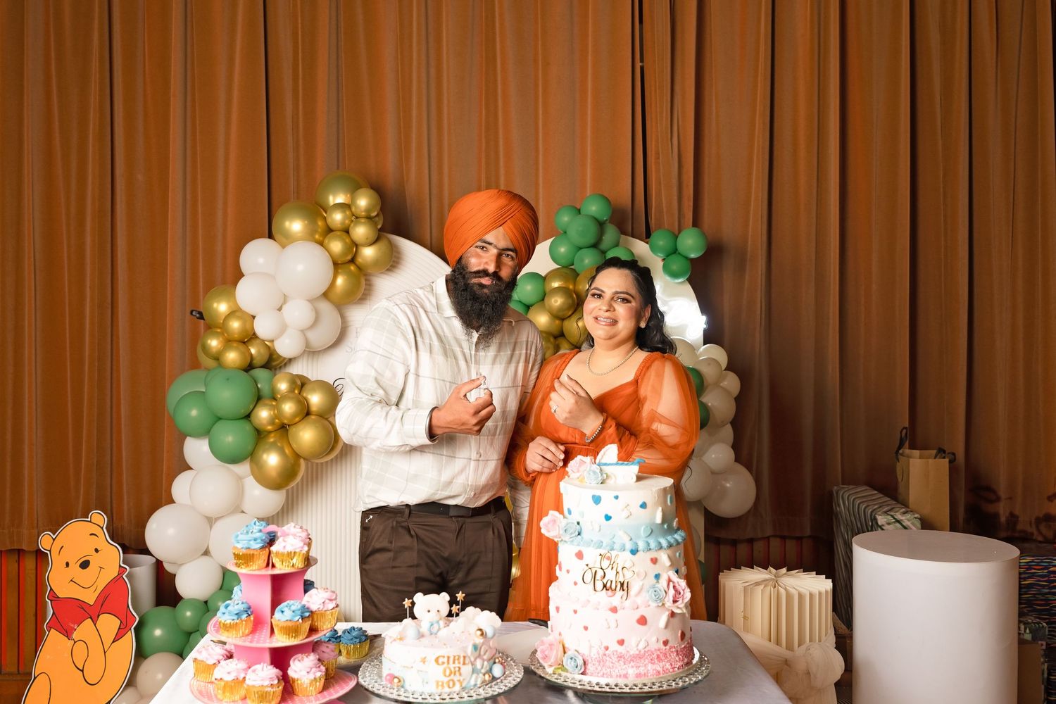 Smiling Sikh couple stands beside baby shower cake and Winnie the Pooh decorations with gold balloon arch.