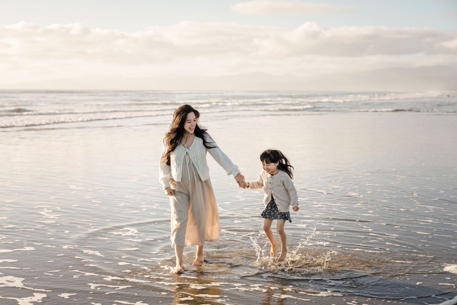A mother and young daughter hold hands while splashing in ocean waves on a sandy beach at sunset.
