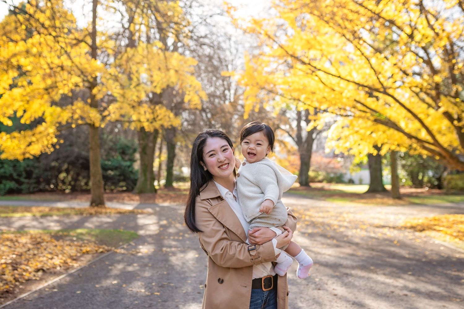A mother and child enjoy a sunny autumn day surrounded by golden yellow trees in a park setting.