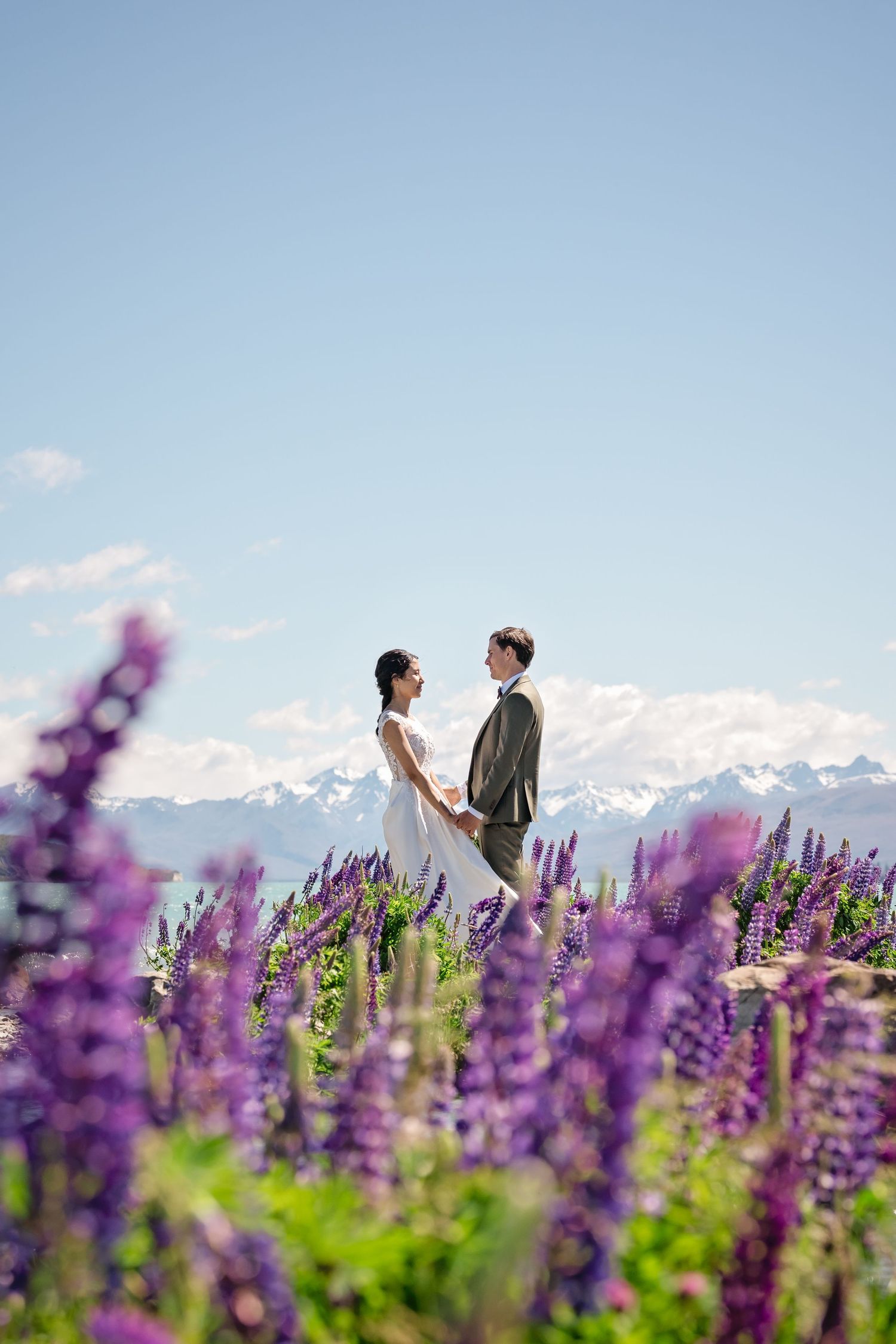 A couple stands together in a vibrant purple lupine field with snow-capped mountains in the background.