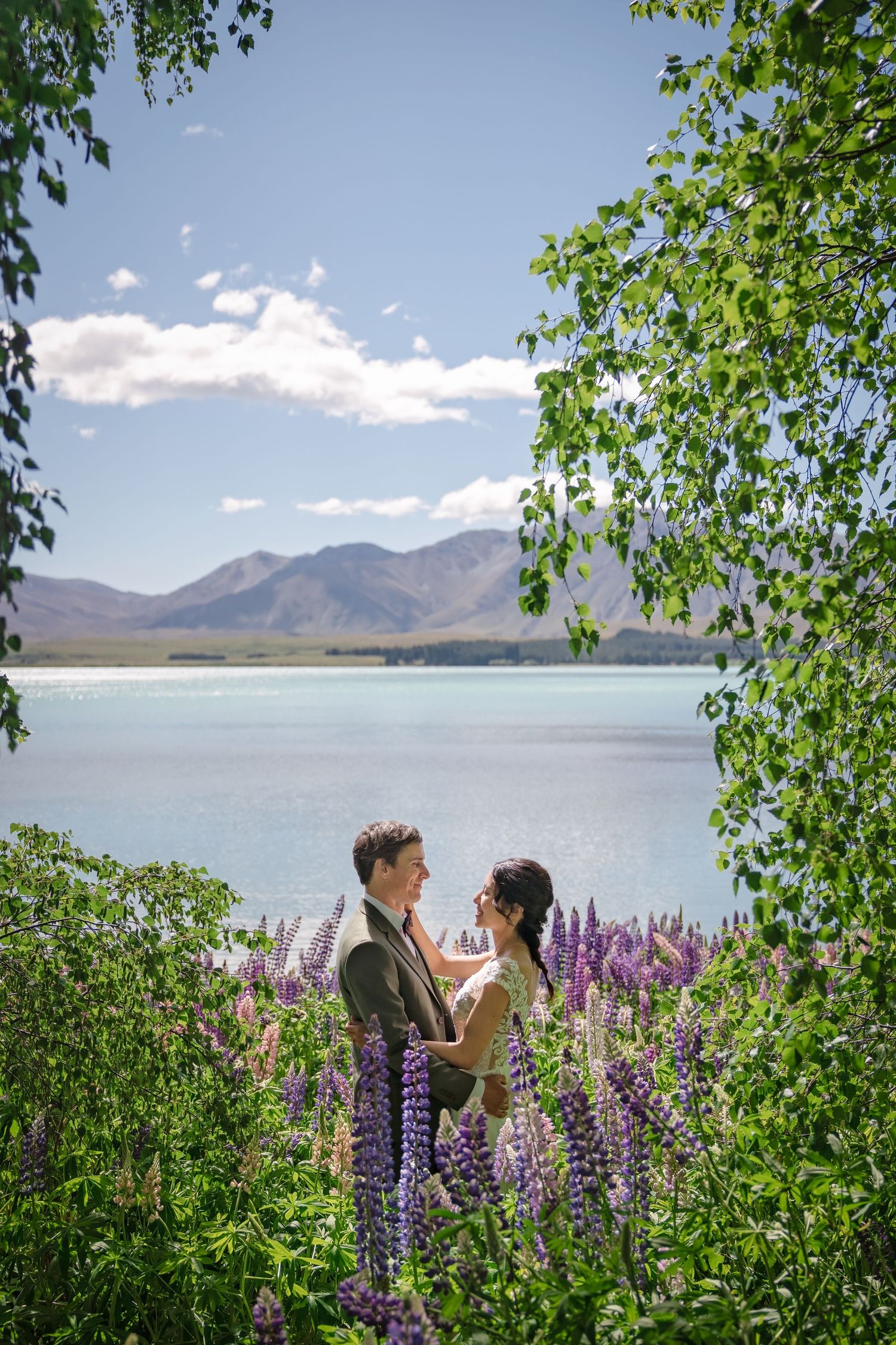 A couple stands together among blooming purple lupines by a serene lake with mountains in the background.