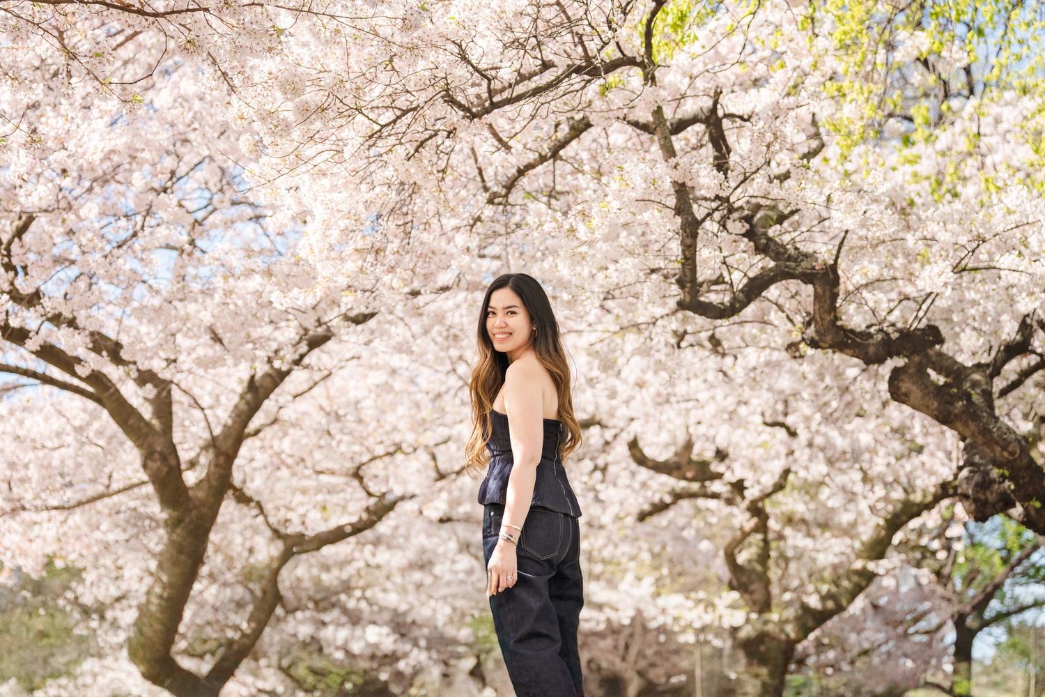 Woman in black outfit posing among blooming cherry blossom trees in spring.