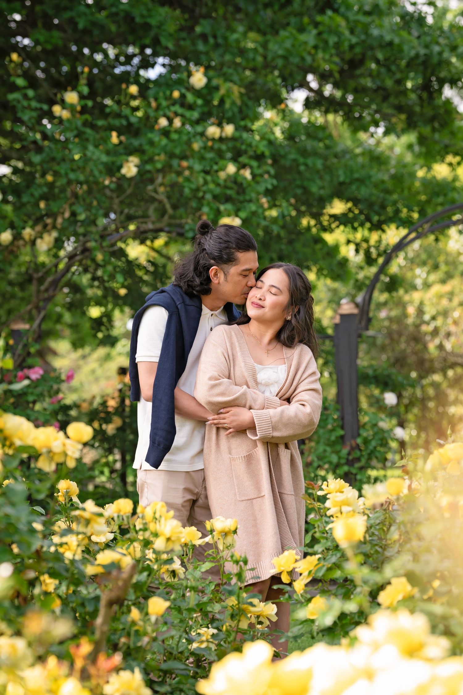 Couple embracing among yellow roses in a lush garden, man kissing woman on cheek.