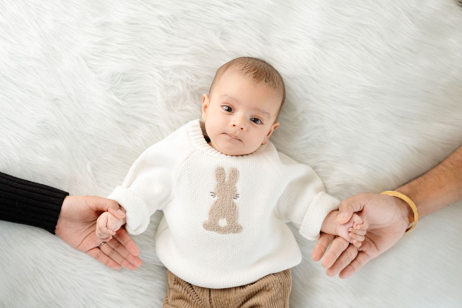 A baby wearing a white bunny sweater lies on fluffy white bedding while holding adult hands on both sides.