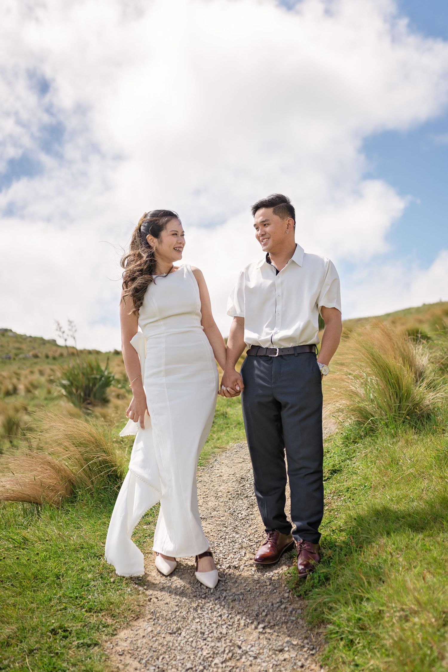 A happy couple holds hands while walking on a gravel path through green hills under a partly cloudy sky.
