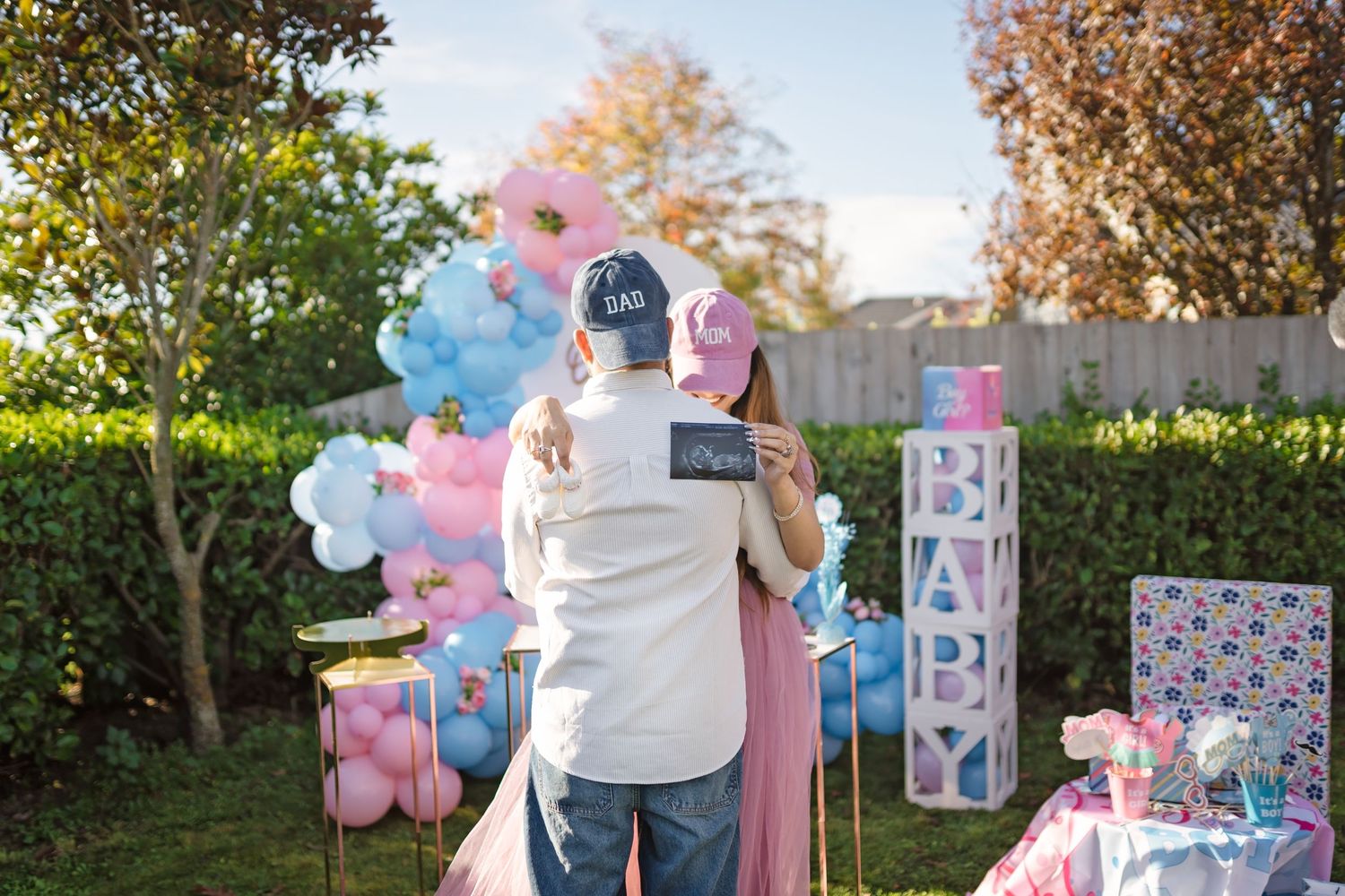 Gender reveal party setup with pink and blue balloon arches, decorative signs and outdoor backyard party decor.