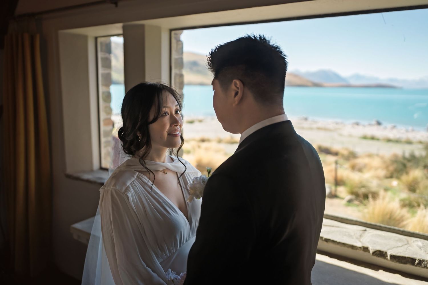 Bride and groom share an intimate moment indoors with a stunning turquoise lake and mountains visible through the window.