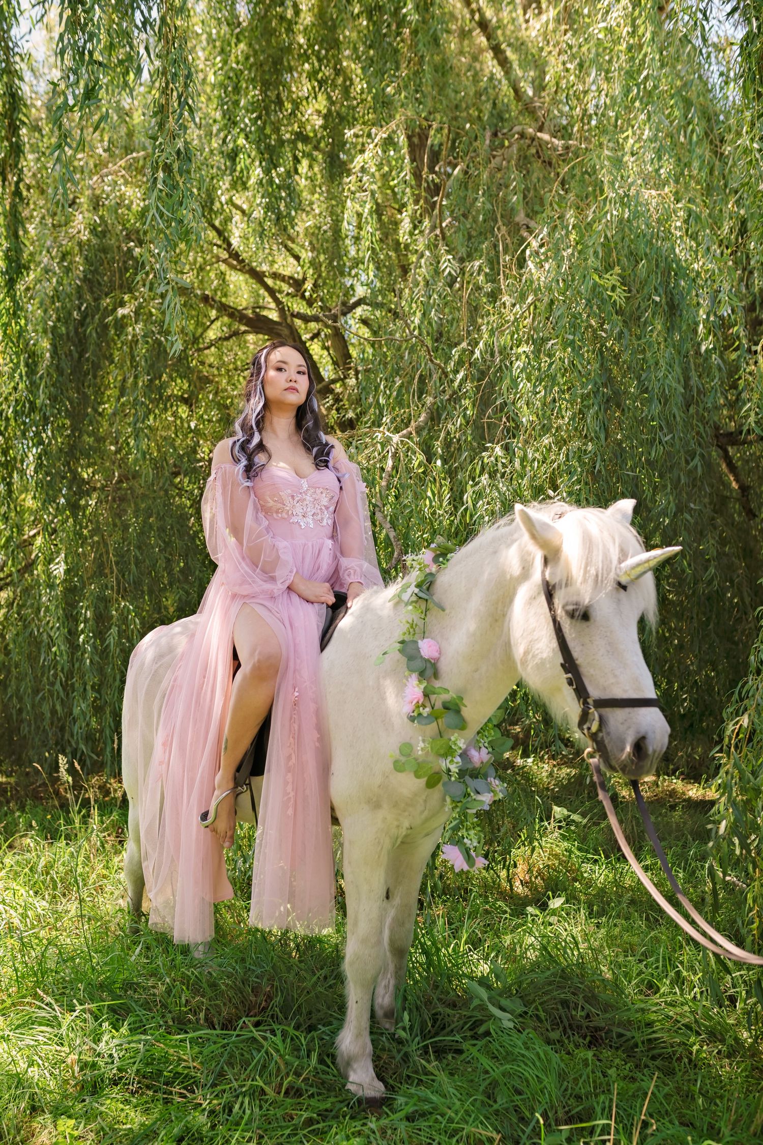 A woman in a pink gown rides a white horse adorned with flowers beneath a weeping willow tree.