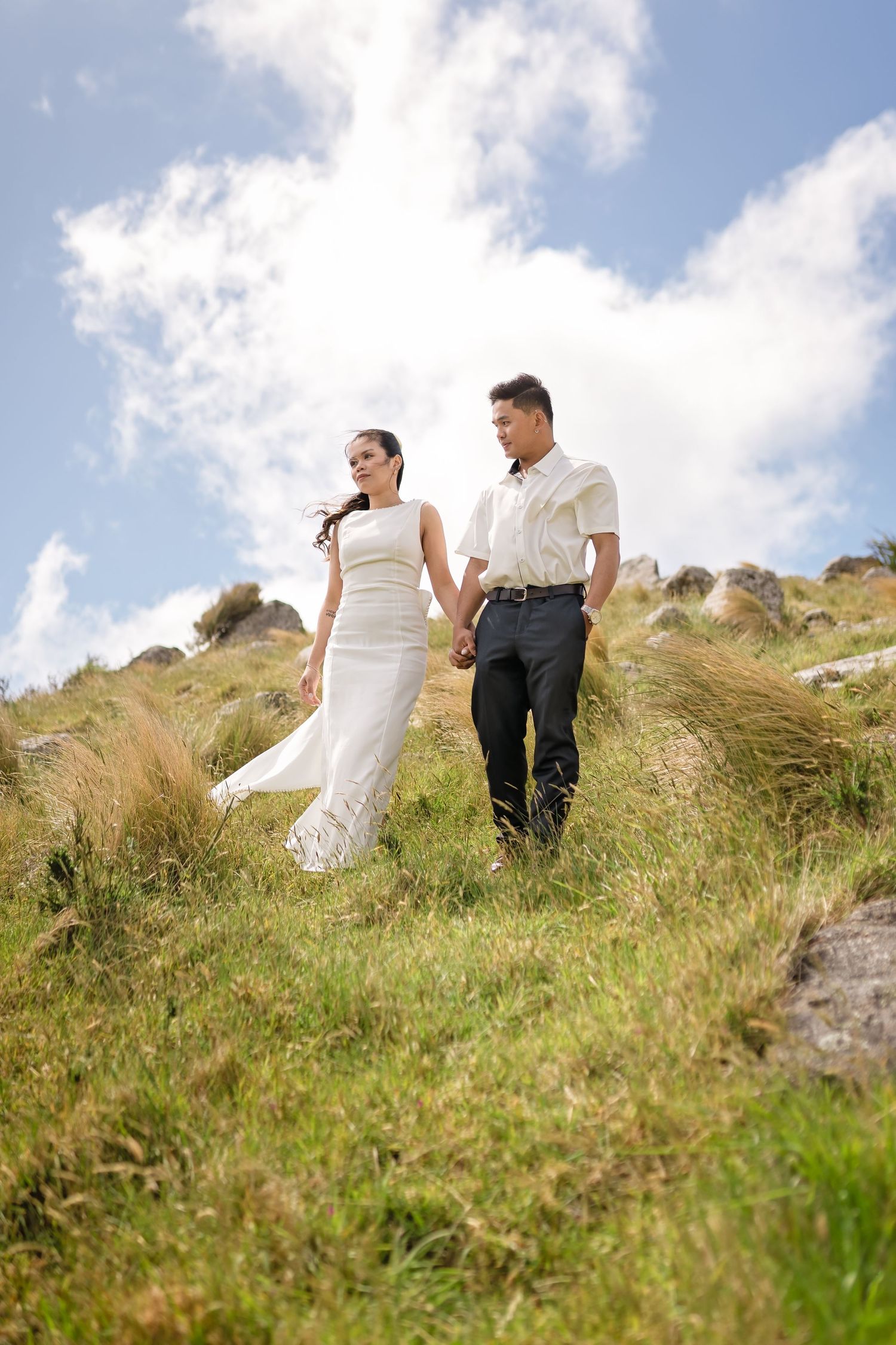 A bride and groom hold hands walking on a grassy hillside under a blue sky with white clouds.