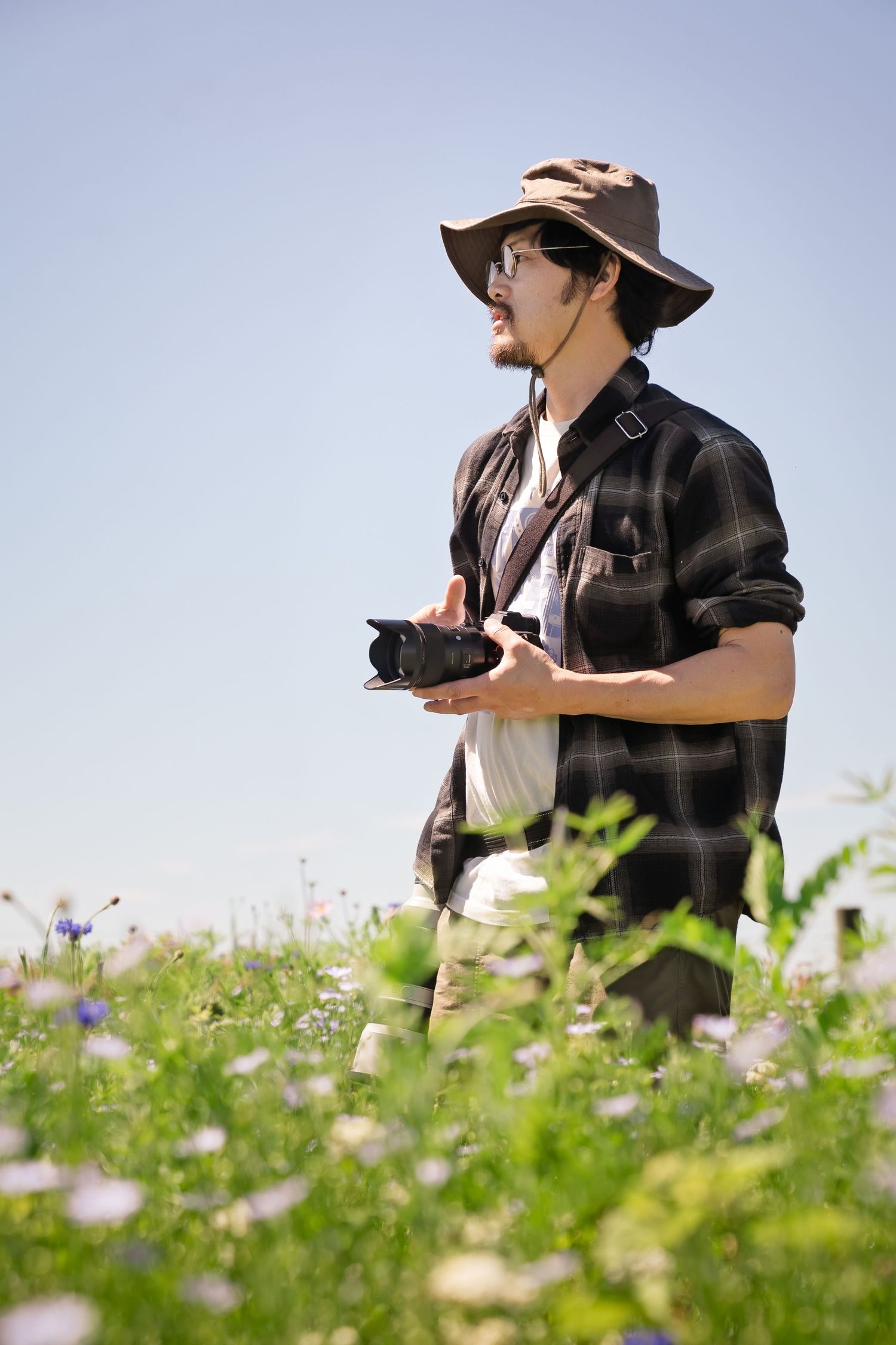 A male photographer in a plaid shirt and bush hat holds a camera while standing in a wildflower meadow under a clear blue sky.