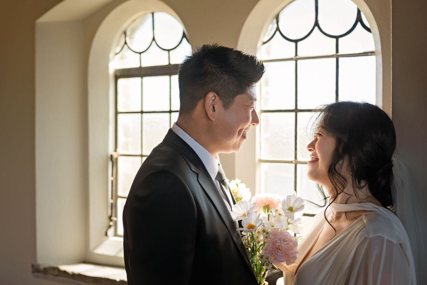 A couple in wedding attire shares a romantic moment by arched windows, bride holding white flowers.
