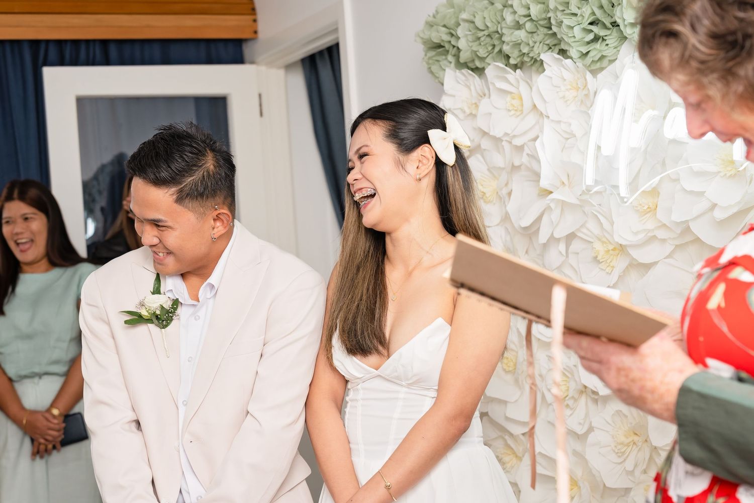 Happy couple laughing during intimate wedding ceremony with floral backdrop and officiant reading vows.