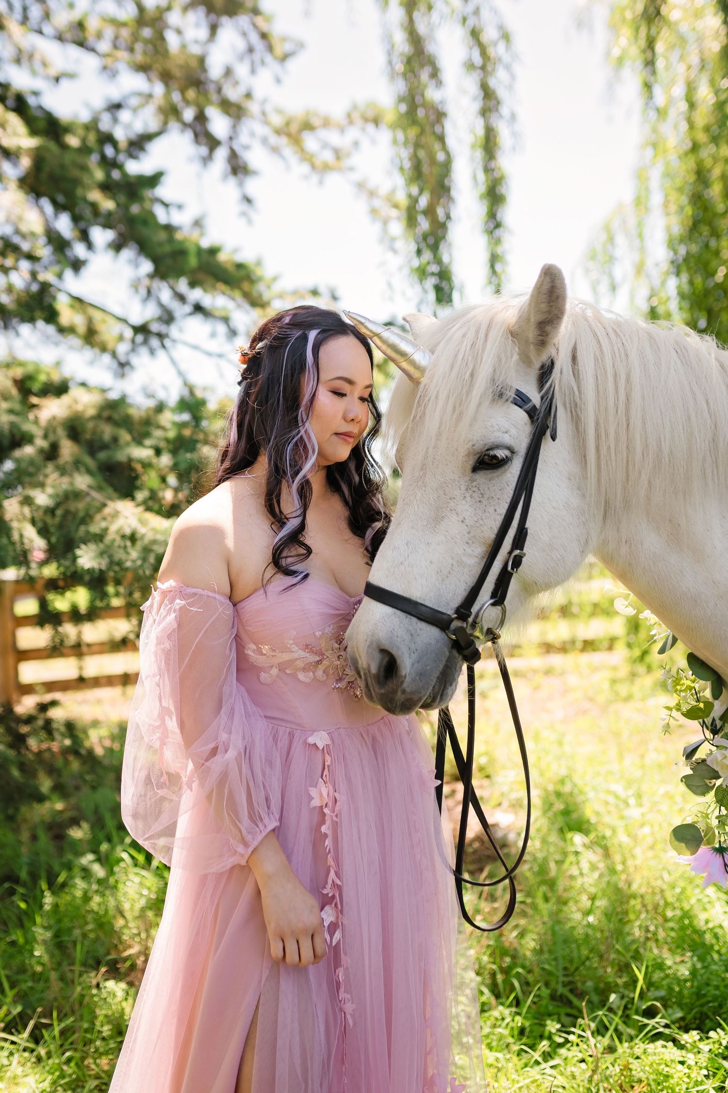 A woman in a pink tulle gown stands beside a white horse in a lush green outdoor setting.