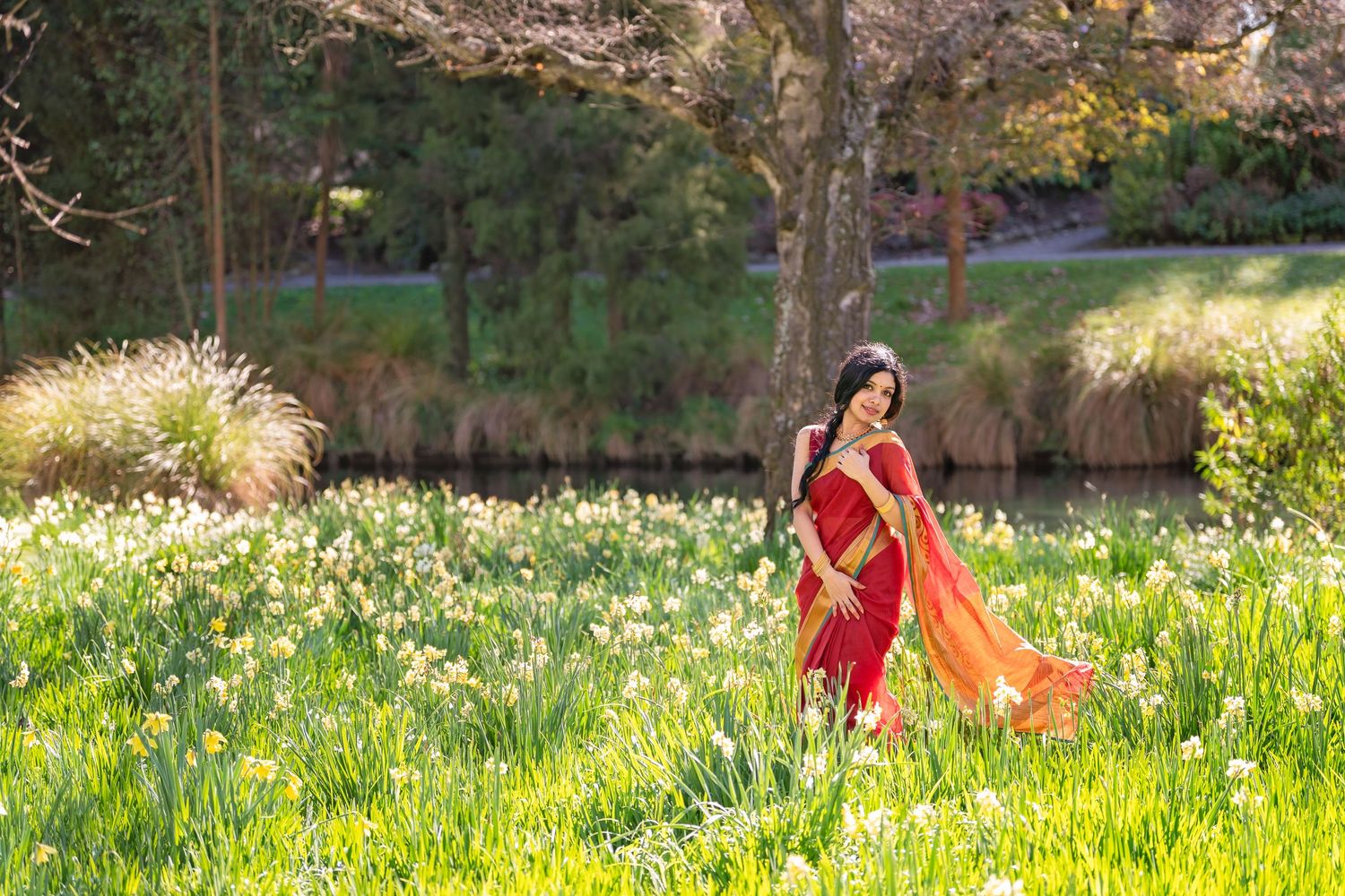 Woman in red saree walking through a field of blooming daffodils beside a serene pond in a lush garden.