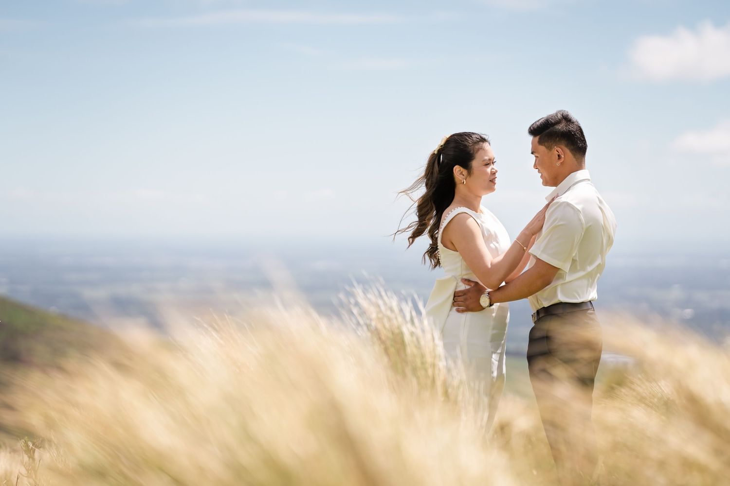 Couple embracing on a scenic hilltop with golden grass and blue sky backdrop.
