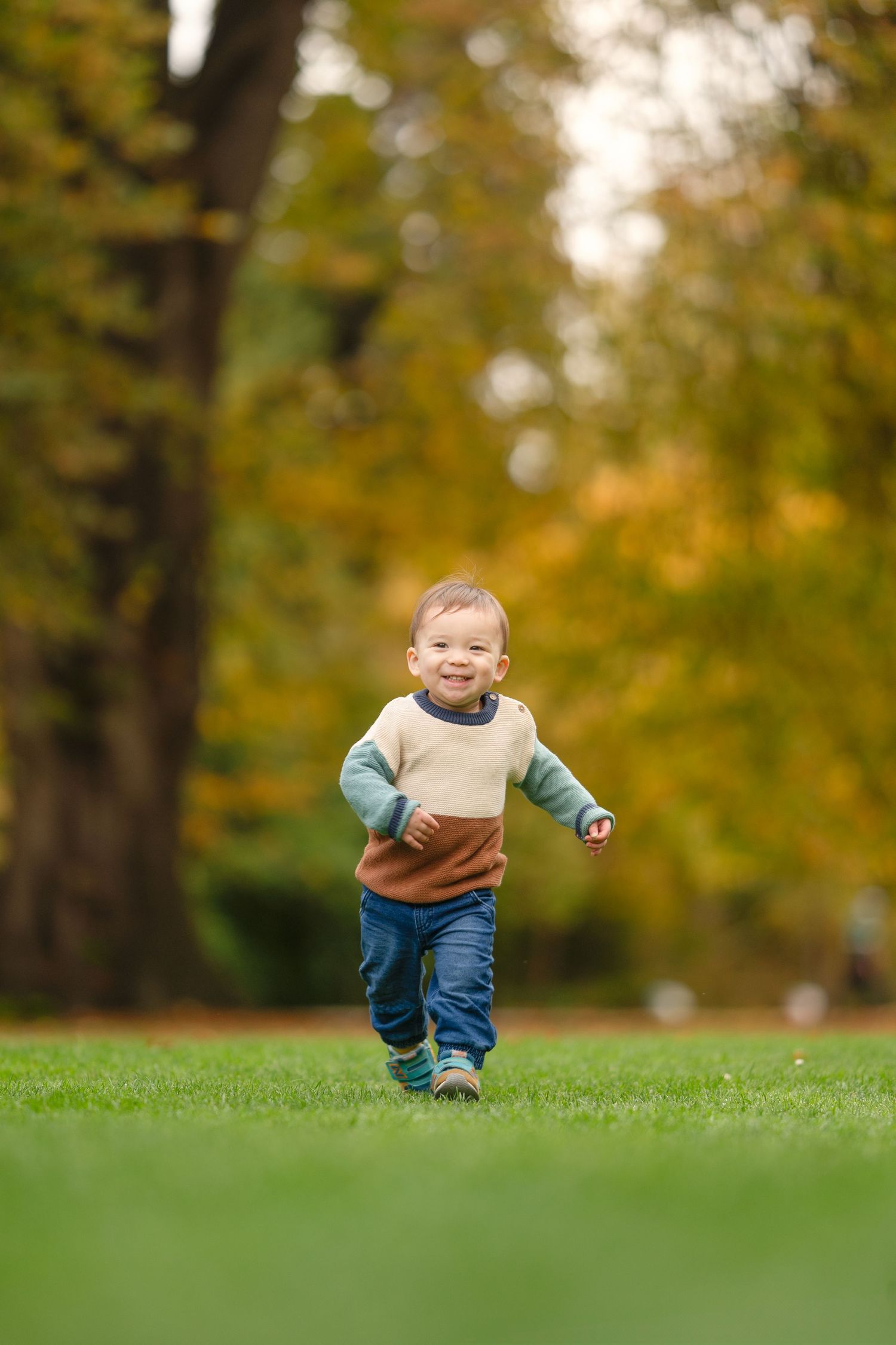 A small child runs joyfully across green grass on an autumn day with golden trees in the background.