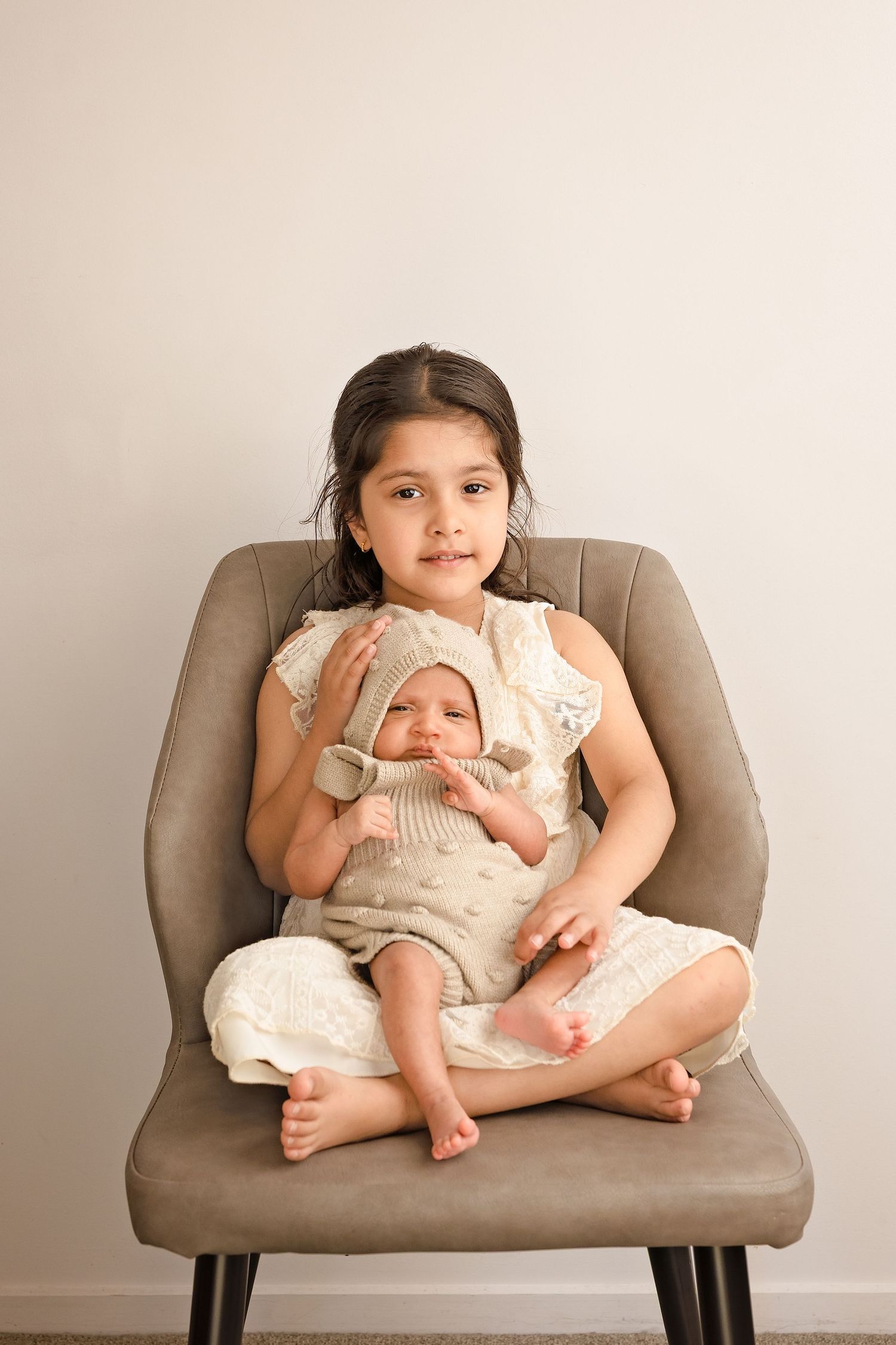 Young girl sitting in gray chair holding newborn baby sibling, both dressed in neutral beige outfits, studio portrait.