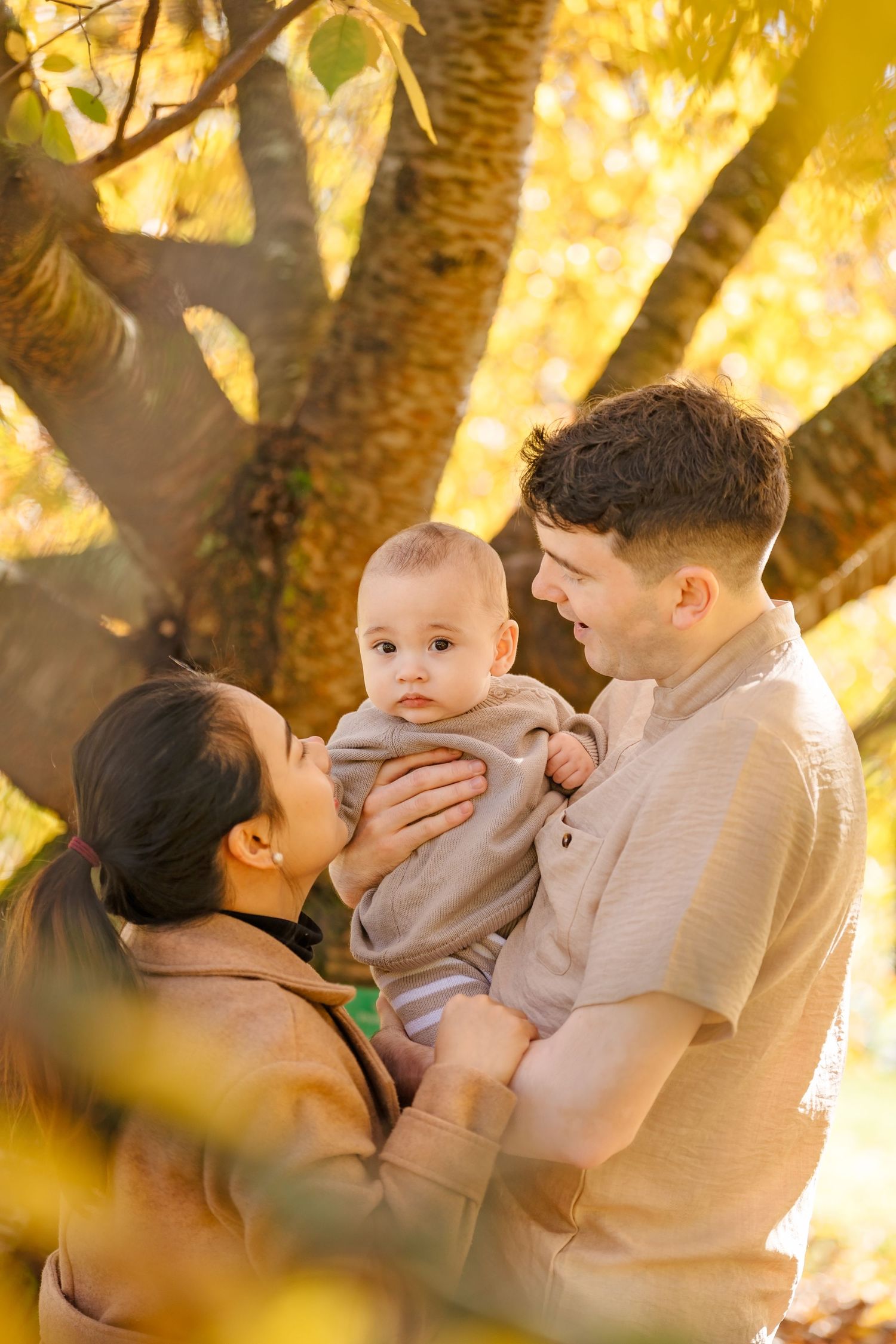 A family shares a tender moment together outdoors in warm autumn light beneath a golden-leafed tree.