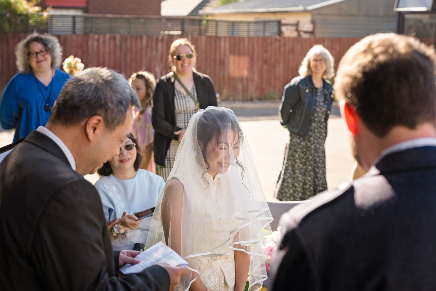 Bride in veil during outdoor wedding ceremony with officiant and guests.