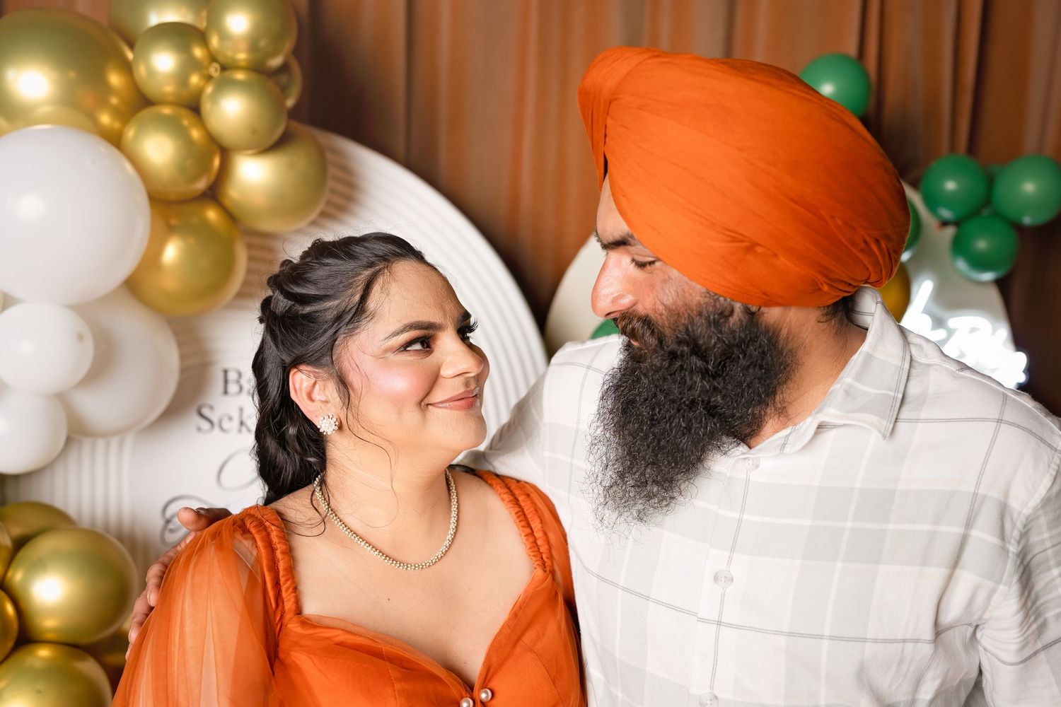 Sikh man in orange turban and beard smiles at woman in orange outfit at decorated baby shower celebration.