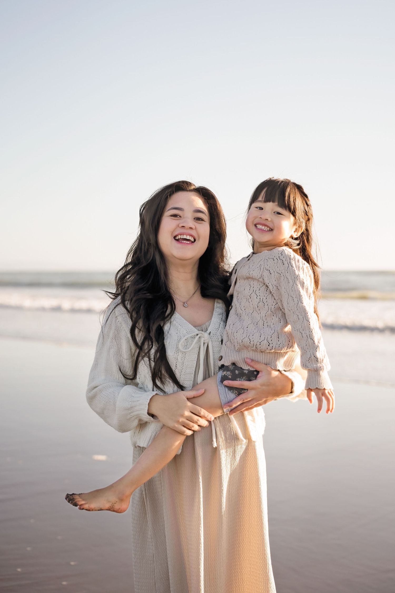 A laughing woman holds a smiling young girl on a sunny beach with ocean waves in the background.