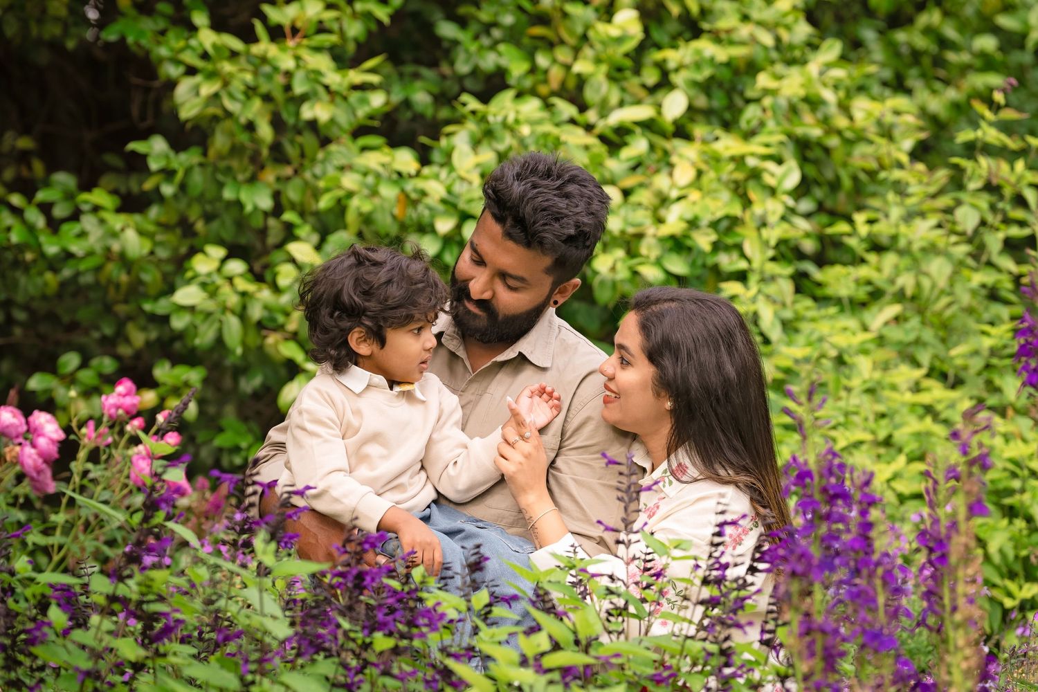 A happy family of three enjoys a sunny day surrounded by colorful purple and pink flowers in a lush garden.