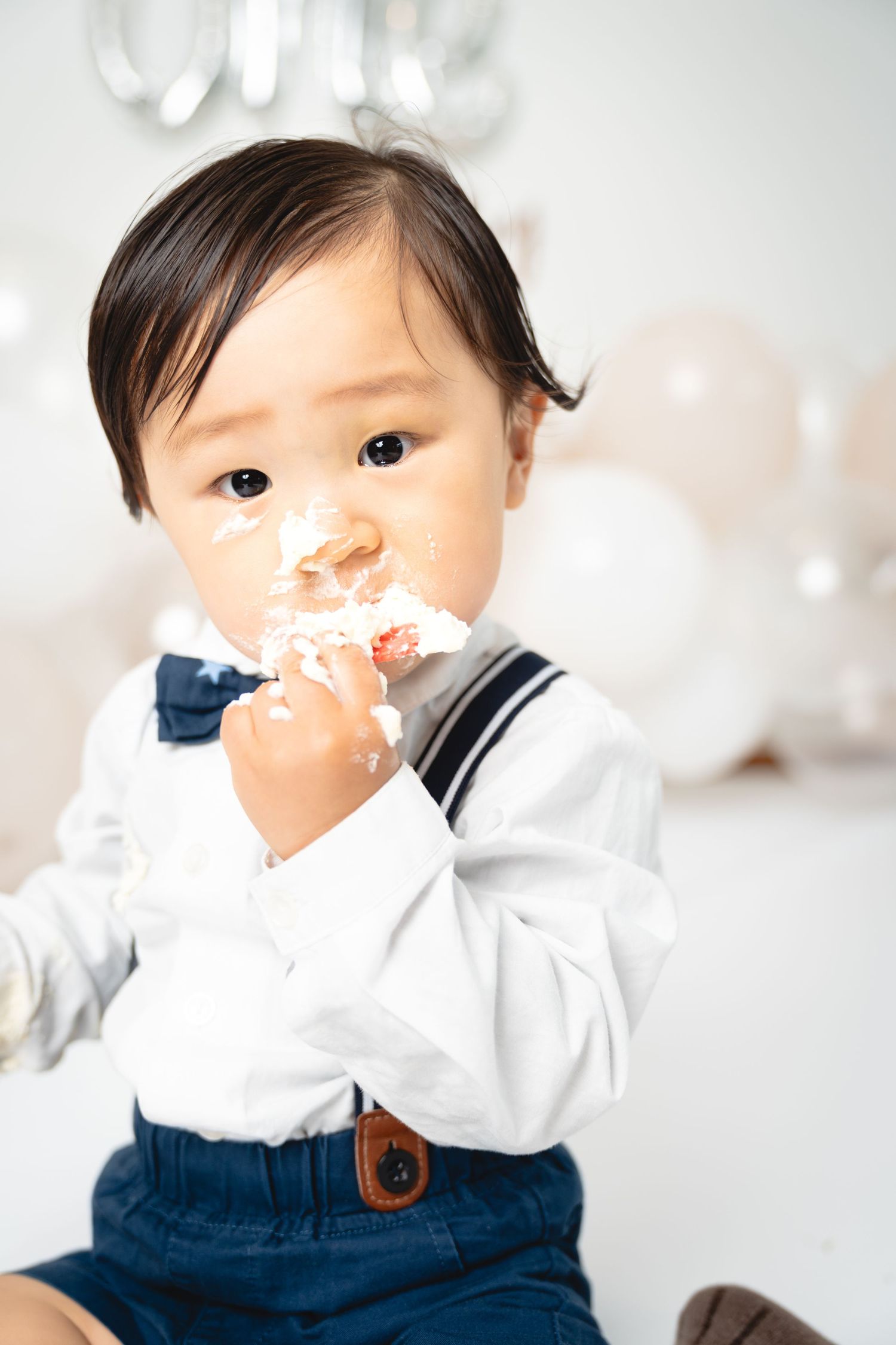 A toddler in suspenders and bow tie enjoys birthday cake with frosting on their face.