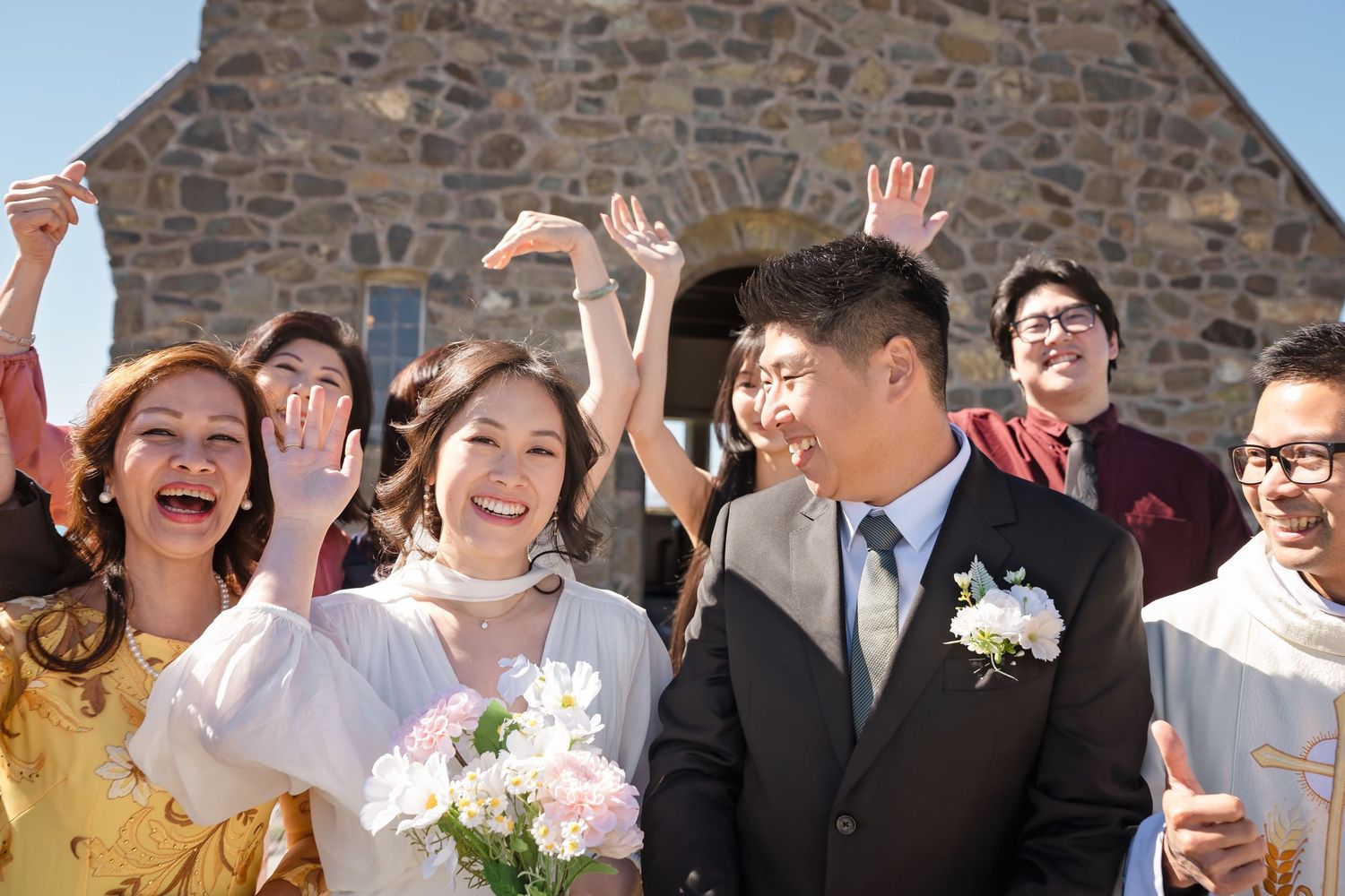 Joyful wedding party celebrates outside a stone church with the happy bride and groom.