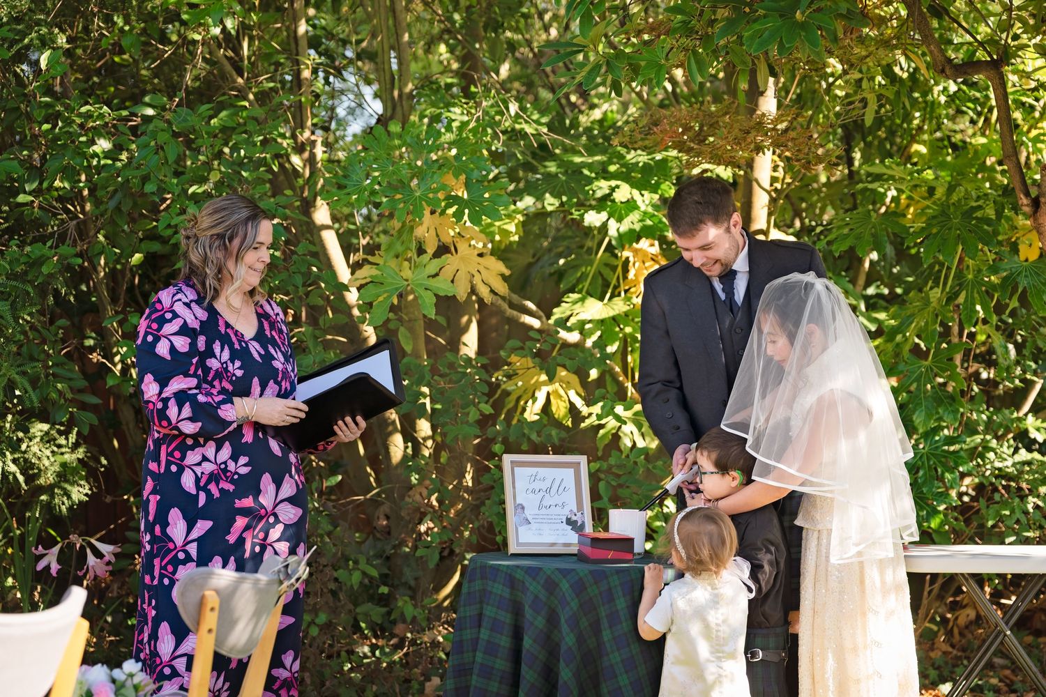 Outdoor wedding ceremony with officiant in floral dress, groom in suit, bride in veil, and flower girl in garden setting.