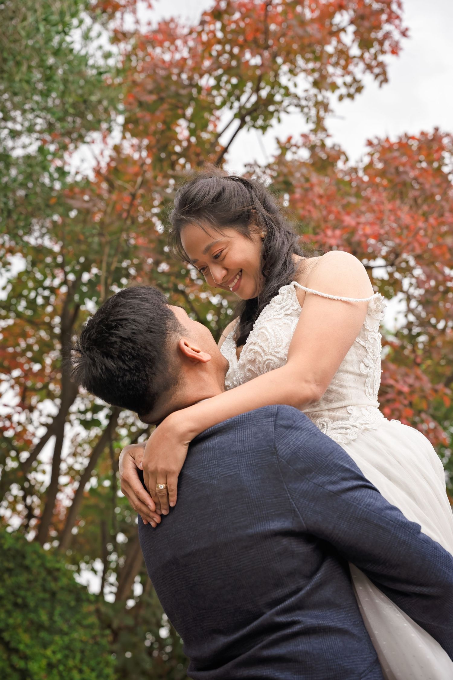 A groom lifts his smiling bride outdoors amid vibrant red and green autumn trees in a romantic wedding moment.