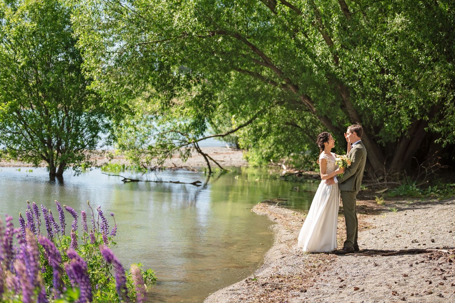 Bride and groom standing by a serene river under lush green trees with purple wildflowers in the foreground.