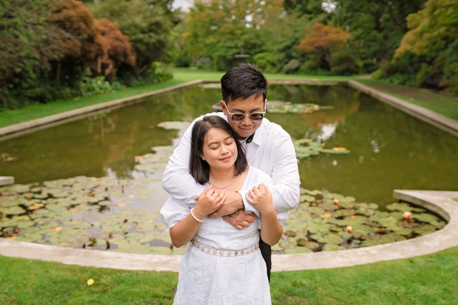 Couple in white outfits embracing by a serene lily pond in a lush green garden setting.