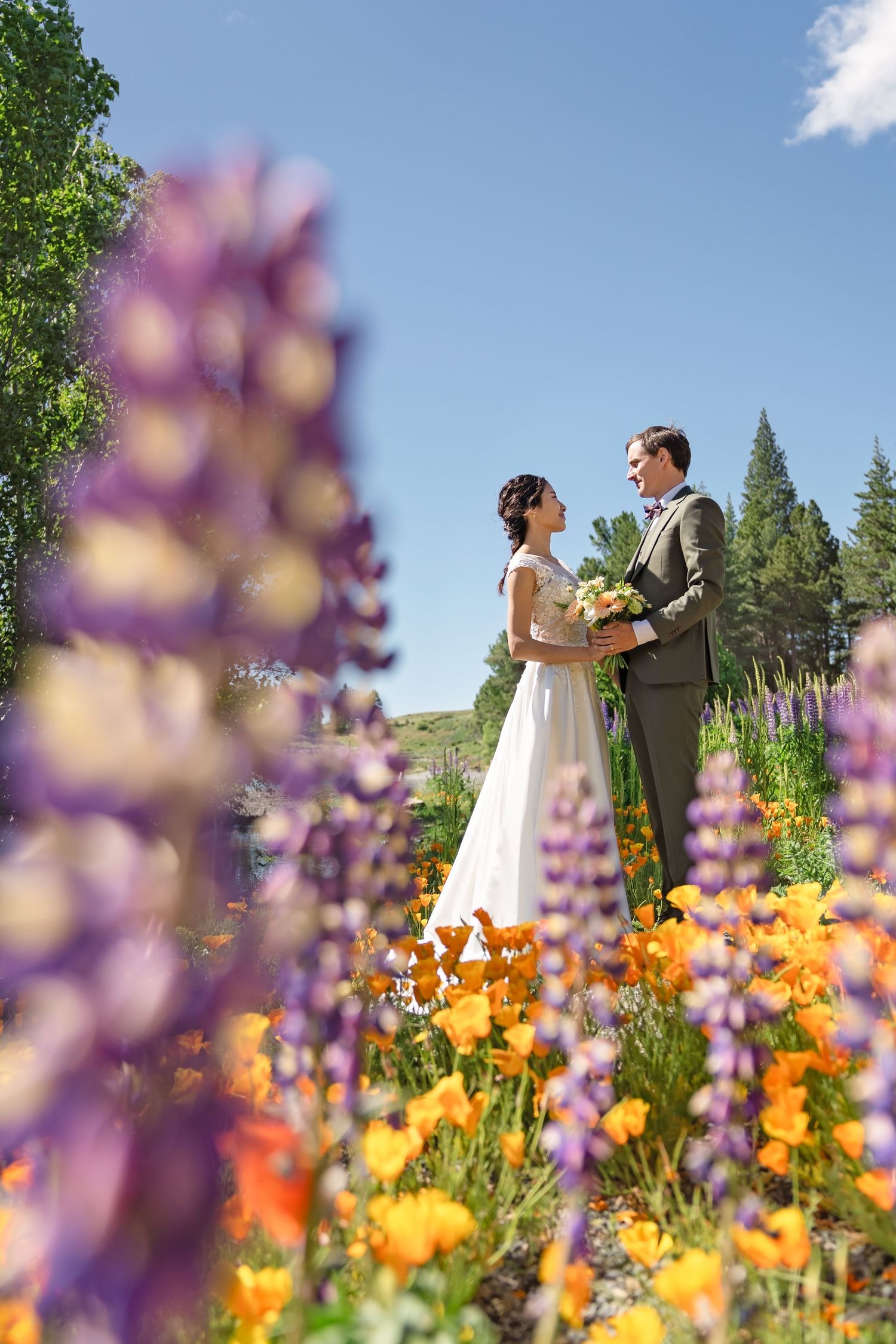 A couple in wedding attire stands together in a meadow filled with vibrant purple lupines and orange wildflowers.