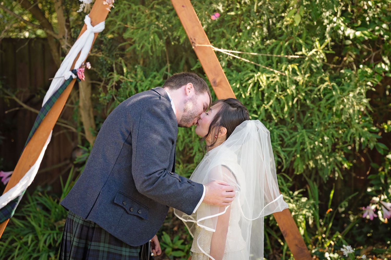Bride and groom share first kiss under wooden arch decorated with ribbons at outdoor garden wedding.