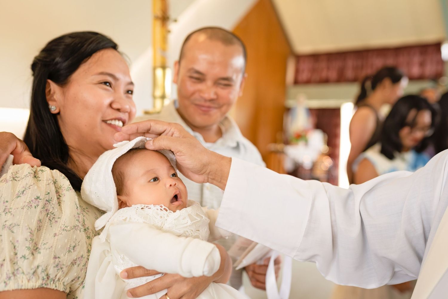 Smiling parents holding baby in white christening gown during baptism ceremony in a church.